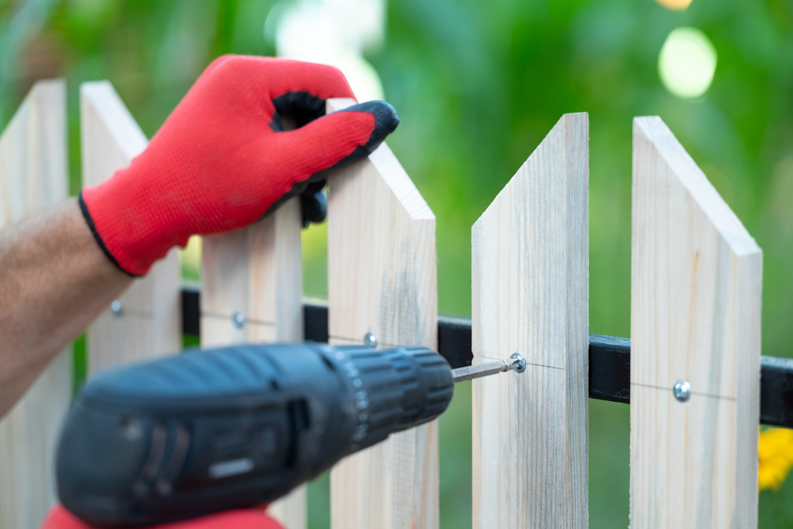 Man hands in working gloves building wooden fence with help of electric screwdriver, close up. Tidying up the home plot yourself, making new fence with own equipment